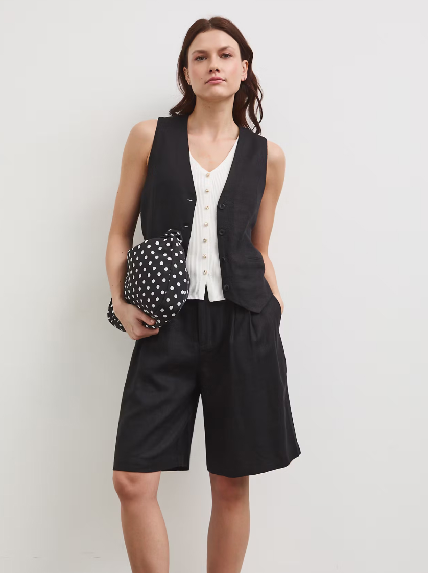 Elegantní letní outfit: žena v černé vestě, světlém topu a volných černých šortkách. Pod paží drží kabelku s velkými bílými puntíky.