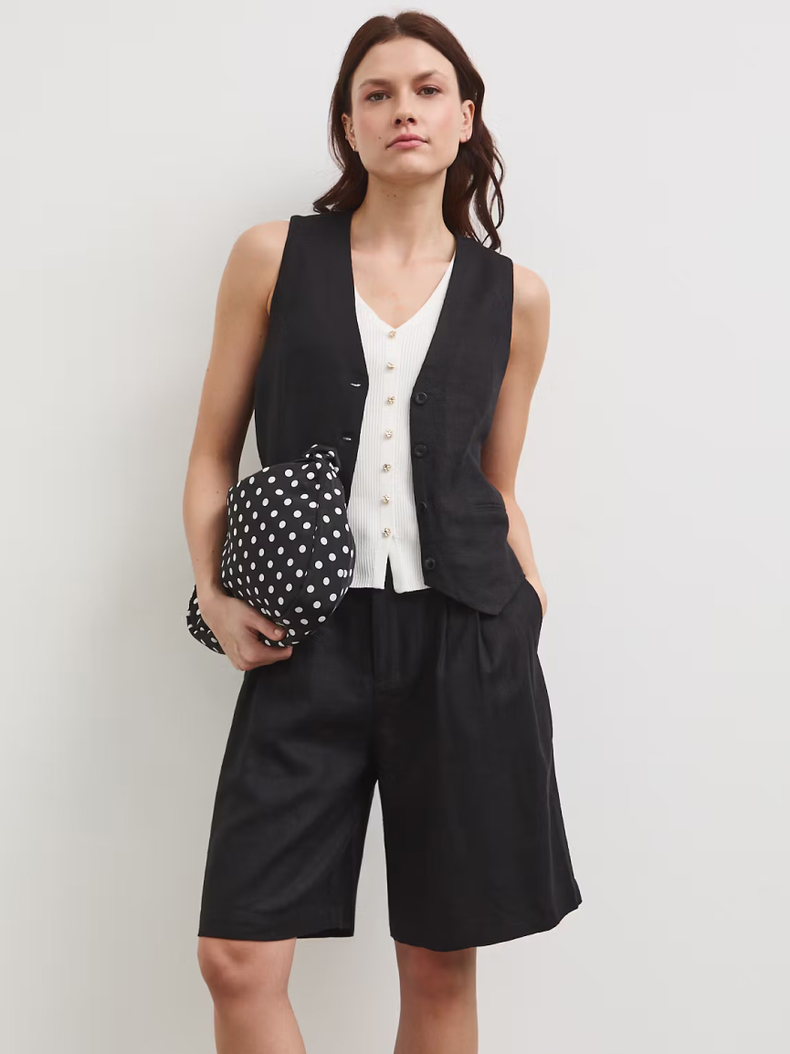 Elegantní letní outfit: žena v černé vestě, světlém topu a volných černých šortkách. Pod paží drží kabelku s velkými bílými puntíky.